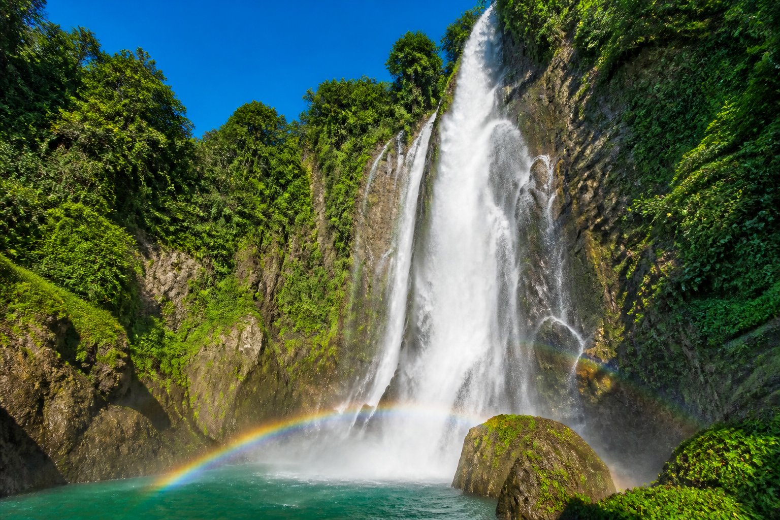 Cachoeira majestosa com água caindo sobre rochas cercadas por vegetação verde e densa em um dia ensolarado.